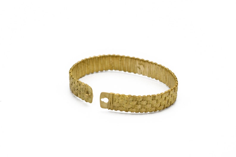 May include: A gold-colored cuff bracelet with a woven texture. The bracelet is open at one end, revealing a clasp. The design features a repeating pattern, and the overall look is vintage.