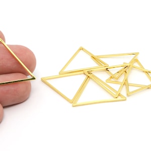 May include: Gold-colored triangle-shaped jewelry findings. Several triangles are scattered on a white surface, with one held by a hand. These geometric shapes are likely used for crafting earrings, necklaces, or other jewelry pieces.