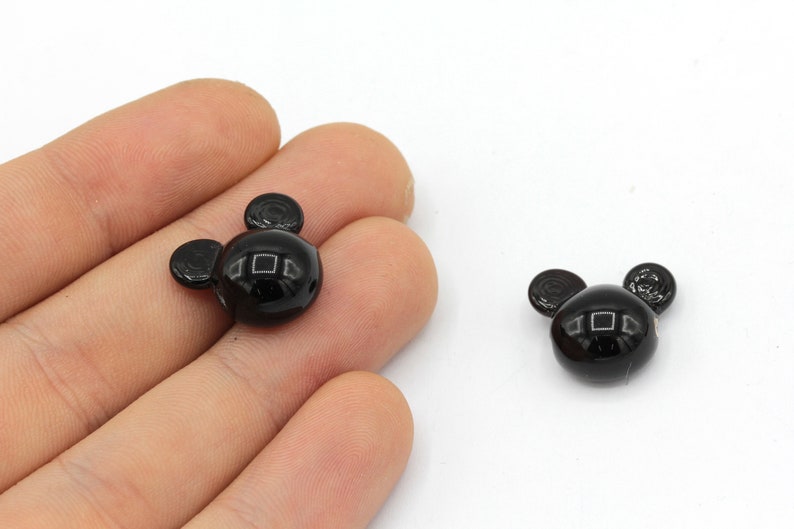 May include: Two glossy black Mickey Mouse-shaped beads. The beads have a round head with two smaller circular ears. The beads are on a white background.