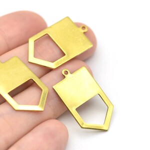 Może przedstawiać: Trzy złote metalowe zawieszki z geometrycznym wzorem. Każda zawieszka ma prostokątną górę z małą pętlą do mocowania do łańcuszka lub sznurka oraz wyciętym sześciokątem.