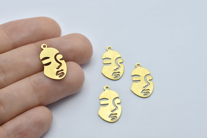 May include: Gold-colored metal face charms with a minimalist design. Each charm features a stylized face with closed eyes and a small loop at the top for attaching to jewelry. The charms are approximately 1 inch in length.