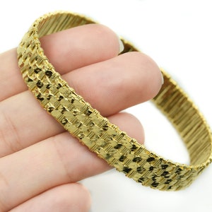May include: A gold-colored bracelet with a woven design. The bracelet is held in a hand against a white background. The bracelet's woven pattern is made up of small, rectangular links.