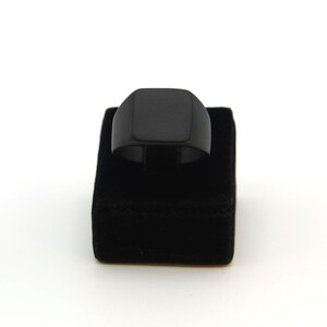 May include: A black signet ring with a square face is displayed on a black velvet ring holder against a white background. The ring has a simple, modern design, and the dark colour provides a bold contrast.