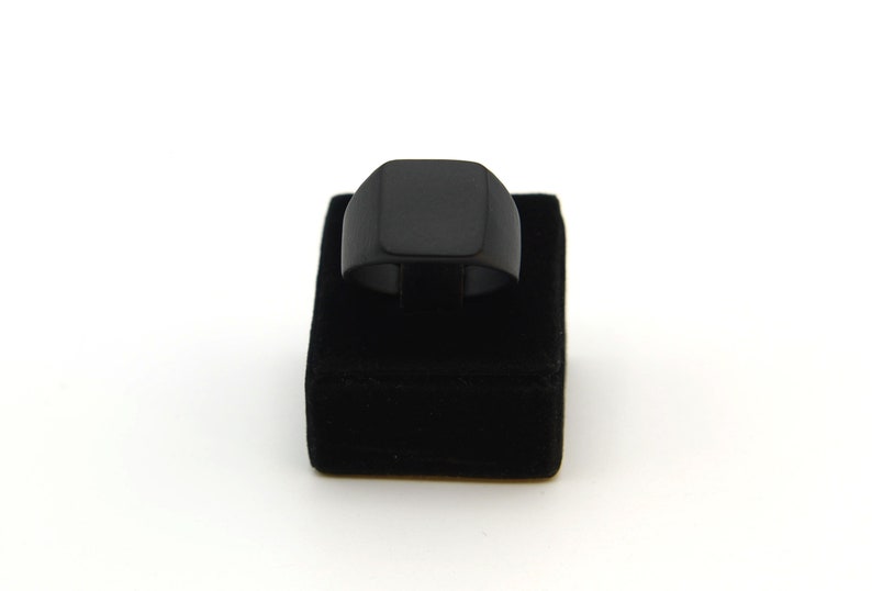 May include: A black signet ring with a square face is displayed on a black velvet ring holder against a white background. The ring has a simple, modern design, and the dark colour provides a bold contrast.