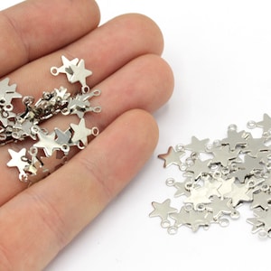 May include: A collection of small, silver-colored star charms with three small loops for attaching to jewelry or other crafts. The charms are scattered on a white surface, with some held in a person's hand.