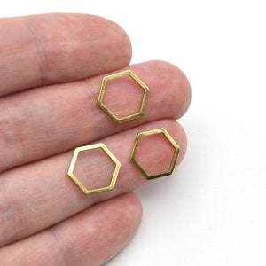 May include: Three small, open-frame, gold-colored hexagon charms are displayed against a white background. The charms are held in a person's hand. These geometric charms are suitable for jewelry making or craft projects.