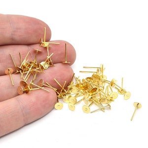 May include: A collection of gold-colored earring posts with flat, round pads and straight posts. The earring findings are scattered on a white surface, ready for jewelry making. The posts are small, with a simple design.