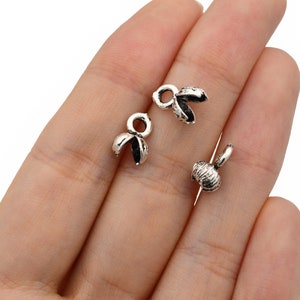 May include: Close-up of several small, silver-colored bead caps with a textured finish. The caps have a petal-like design and are open, revealing the interior. One cap is attached to a needle.