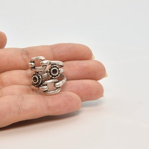 May include: A silver-toned ring with a unique design. The ring features a series of interconnected links and small, circular embellishments with dark centers. The ring is held in a hand against a white background.