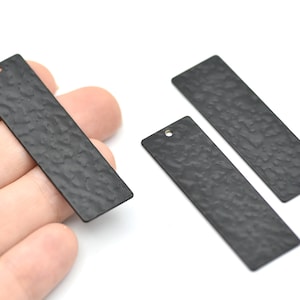 May include: Three rectangular, black metal jewellery tags with a hammered texture. One tag is held in a hand. Each tag has a small hole at one end for attaching to jewellery. The tags are on a white background.
