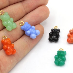 May include: A collection of colorful gummy bear charms in green, blue, black, orange, and red. Each charm has a gold-colored loop for attaching to jewelry or other crafts. The charms are small and have a glossy finish.