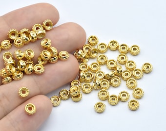 May include: A collection of small, gold-colored metal beads. Each bead has a cylindrical shape with a textured, decorative pattern around the edges. The beads are scattered on a white surface, with some held in a hand.