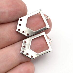 Może przedstawiać: Dwa srebrne metalowe elementy biżuteryjne. Każdy element ma geometryczny kształt z centralnym otwartym obszarem i wieloma małymi otworami. Jeden ma pętlę do mocowania. Elementy znajdują się na białym tle.