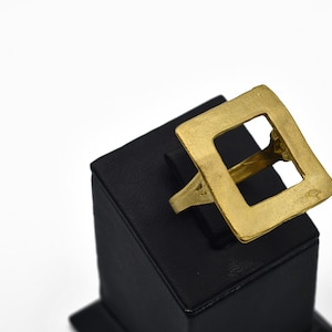 Op de afbeelding: Goudkleurige ring met een vierkant ontwerp. De ring wordt gepresenteerd op een zwarte standaard tegen een witte achtergrond. Het vierkant is in het midden open.