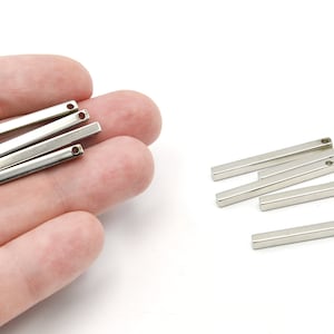 May include: Several silver-colored rectangular metal bars with a small hole at one end. Some bars are held in a hand, while others are scattered on a white surface. These components are likely for jewelry making or crafts.
