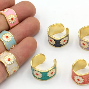 May include: Adjustable rings with a gold band and colorful enamel daisy design. The rings feature a variety of colors, including pink, light blue, cream, black, purple, teal, and red. The daisy flowers have white petals and a red center.