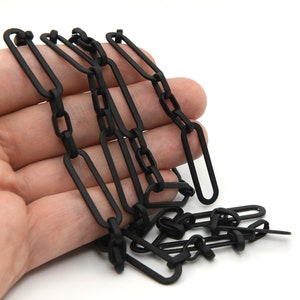May include: Black chain with elongated oval links and smaller connecting links. The chain is held in a hand against a white background. The chain is likely used for jewelry or craft projects.