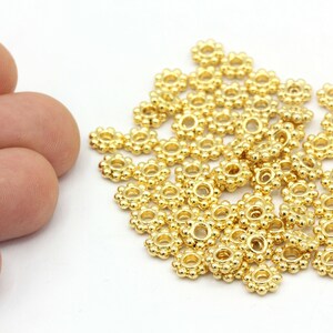 Peut inclure: Gros plan sur des perles dor&eacute;es en forme de fleur. Chaque perle a un trou central entour&eacute; de petits accents ronds. Quelques perles sont tenues dans une main, avec un plus grand tas de perles en arri&egrave;re-plan. Les perles sont probablement pour la fabrication de bijoux.