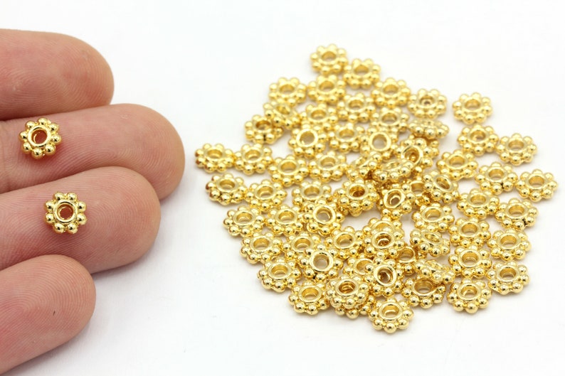 Peut inclure: Gros plan sur des perles dor&eacute;es en forme de fleur. Chaque perle a un trou central entour&eacute; de petits accents ronds. Quelques perles sont tenues dans une main, avec un plus grand tas de perles en arri&egrave;re-plan. Les perles sont probablement pour la fabrication de bijoux.