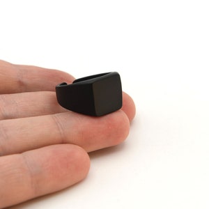 Op de afbeelding: Een matzwarte ring met een vierkante voorkant. De ring wordt getoond op de hand van een persoon tegen een witte achtergrond. Het ontwerp van de ring is eenvoudig en modern, geschikt voor dagelijks gebruik.