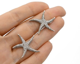 Op de afbeelding: Twee zilveren oorbellen in de vorm van zeesterren, elk bezet met kleine, heldere kristallen. De oorbellen worden in een hand getoond tegen een witte achtergrond. Het zeesterontwerp is een populair motief voor sieraden met strandthema.