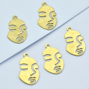 May include: Five brass-colored face pendants with a minimalist design. Each pendant features a stylized face with cut-out details for the eyes, nose, and mouth. The pendants have a small loop at the top for attaching to jewelry.