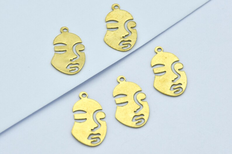 May include: Five brass-colored face pendants with a minimalist design. Each pendant features a stylized face with cut-out details for the eyes, nose, and mouth. The pendants have a small loop at the top for attaching to jewelry.
