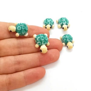 May include: A collection of small glass turtle figurines. Each turtle has a teal shell with white circular patterns, cream-coloured head and limbs, and black eyes. The turtles are arranged on a white background.