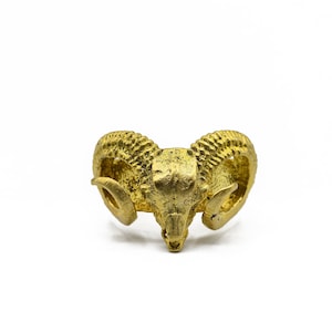 May include: A gold-colored ring shaped like a ram's head, featuring detailed horns and facial features. The ring is set against a white background, highlighting its metallic texture and design.