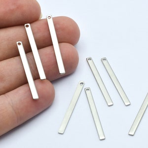 Op de afbeelding: Een verzameling zilverkleurige, rechthoekige metalen staafbedels. Elke bedel heeft een klein gaatje aan de bovenkant. De bedels worden tegen een witte achtergrond getoond.