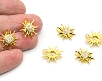 Può includere: Ciondoli a forma di sole dorato con un cerchio centrale di piccoli cristalli trasparenti. Il design a forma di sole presenta raggi appuntiti che si estendono verso l'esterno. Diversi ciondoli sono visibili, alcuni tenuti in mano, su sfondo bianco.
