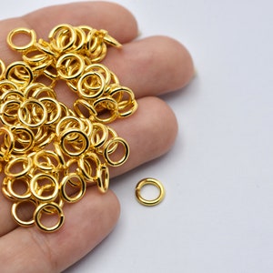 May include: A collection of small, shiny gold-colored jump rings. These open rings are used for jewelry making and other crafts. The rings are scattered across a light background.