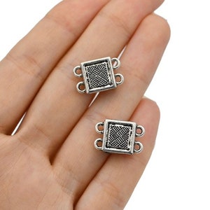 May include: Two small, square, silver-colored metal jewelry connectors. Each connector has a woven pattern in the center and two loops on each side. The connectors are held in a person's hand.