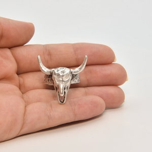 May include: A silver-colored ring featuring a detailed bull skull design. The ring's skull has prominent horns and a textured surface, giving it a rustic, Western-inspired aesthetic. The ring is held in a hand, showcasing its size and design.