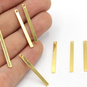 May include: Several gold-colored rectangular metal bars with a small hole at one end. The bars are arranged on a white surface, some held by a hand. These are likely jewelry-making components or decorative elements.