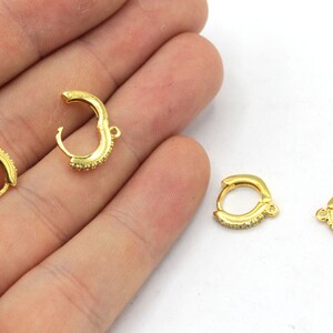 May include: Gold-colored hoop earrings with small, clear stones set along the outer curve. The earrings are shown in an open and closed position, with a small loop for attachment.
