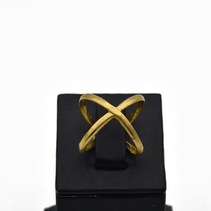 May include: A gold-colored ring with an overlapping X design. The ring is displayed on a black stand against a white background. The ring's design features two curved bands that intersect in the center.