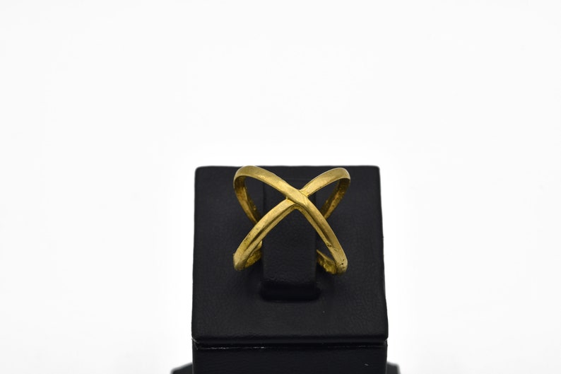 May include: A gold-colored ring with an overlapping X design. The ring is displayed on a black stand against a white background. The ring's design features two curved bands that intersect in the center.