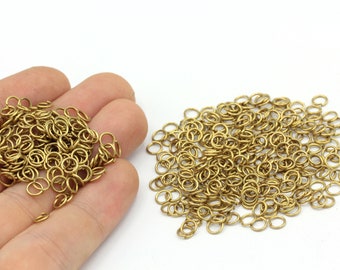Puede incluir: Una colección de pequeños anillos de salto de color latón, dispersos sobre una superficie blanca. Algunos anillos se sostienen en una mano, mientras que otros están extendidos. Estos componentes para la fabricación de joyas son ideales para manualidades.