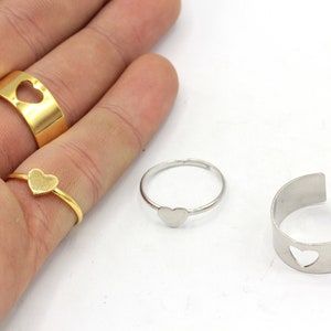 May include: A collection of rings featuring heart designs. Includes a gold band with a heart cutout, a delicate gold ring with a heart, a silver ring with a heart, and a silver adjustable ring with a heart cutout.