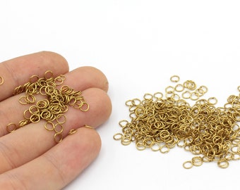 Puede incluir: Una colección de pequeños anillos de salto de color latón esparcidos sobre una superficie blanca. Algunos anillos se sostienen en una mano. Estos anillos abiertos se utilizan para la fabricación de joyas y otras manualidades. Los anillos tienen aproximadamente 0,5 cm de diámetro.
