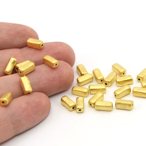 May include: A collection of gold-colored, rectangular beads with rounded edges and small holes. The beads are scattered on a white surface, some held in a hand. These beads are likely used for jewelry making or crafting.