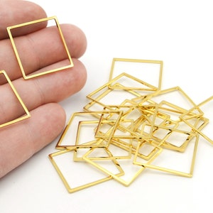 May include: A collection of gold-colored, open-frame square charms. Some are held in a hand, while others are scattered on a white surface. These small, geometric shapes are likely used for jewelry making or crafting.