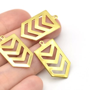 May include: Three gold-colored metal pendants with a geometric chevron design. Each pendant has a rectangular shape with a small loop at the top for attaching to a chain or other jewelry component. The pendants are held in a hand.
