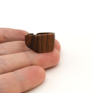 May include: A wooden ring with a geometric design. The ring is made of dark brown wood with visible grain patterns. The ring is square-shaped with a cut-out section on the top. The ring is on a white background.