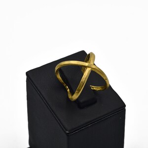 May include: A gold-colored ring with an overlapping X design. The ring is displayed on a black square stand against a white background. The ring's design features two bands that cross each other.