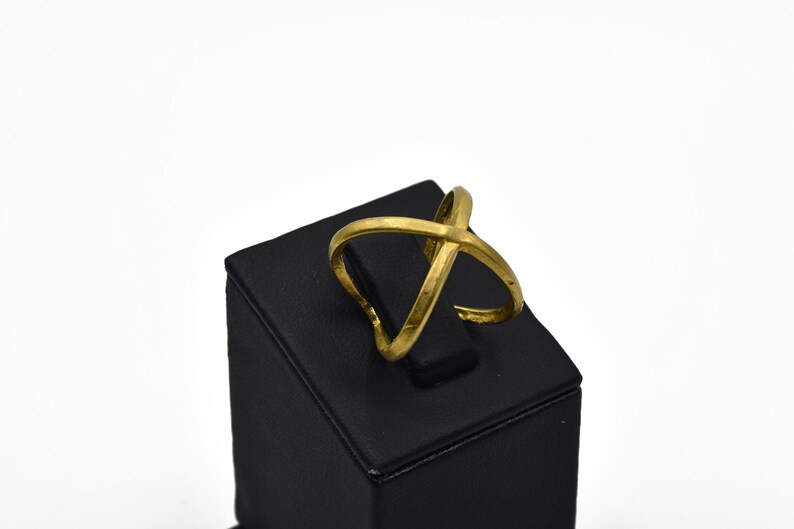 May include: A gold-colored ring with an overlapping X design. The ring is displayed on a black square stand against a white background. The ring's design features two bands that cross each other.