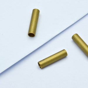 May include: Three small, cylindrical brass tubes are arranged on a white surface. Each tube has a hollow interior and a smooth, metallic finish. The tubes are evenly spaced, suggesting a potential for jewelry making or crafting.