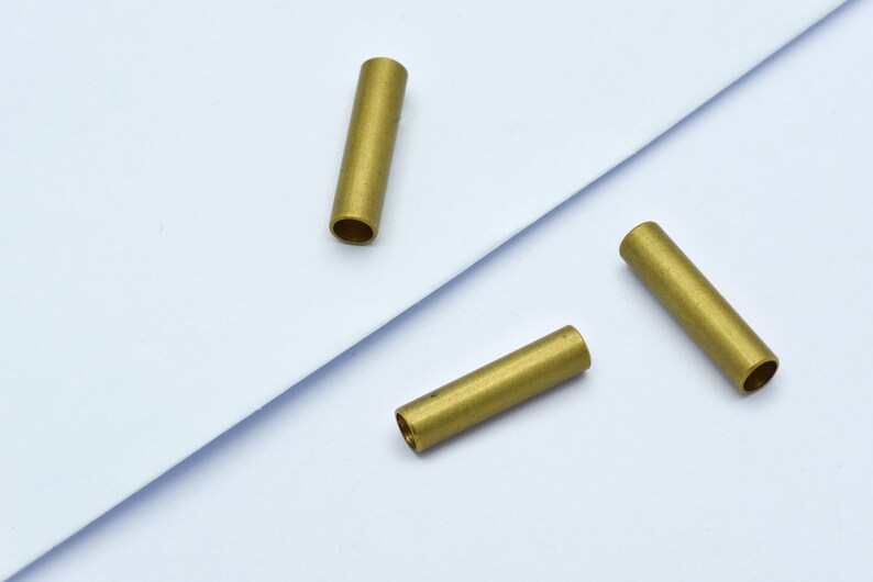 May include: Three small, cylindrical brass tubes are arranged on a white surface. Each tube has a hollow interior and a smooth, metallic finish. The tubes are evenly spaced, suggesting a potential for jewelry making or crafting.
