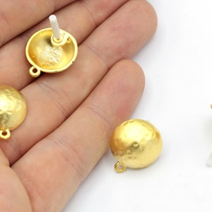 May include: Gold-colored, hammered-texture stud earrings. The earrings have a half-dome shape with a small loop at the base. One earring post is visible. The earrings are shown against a white background.